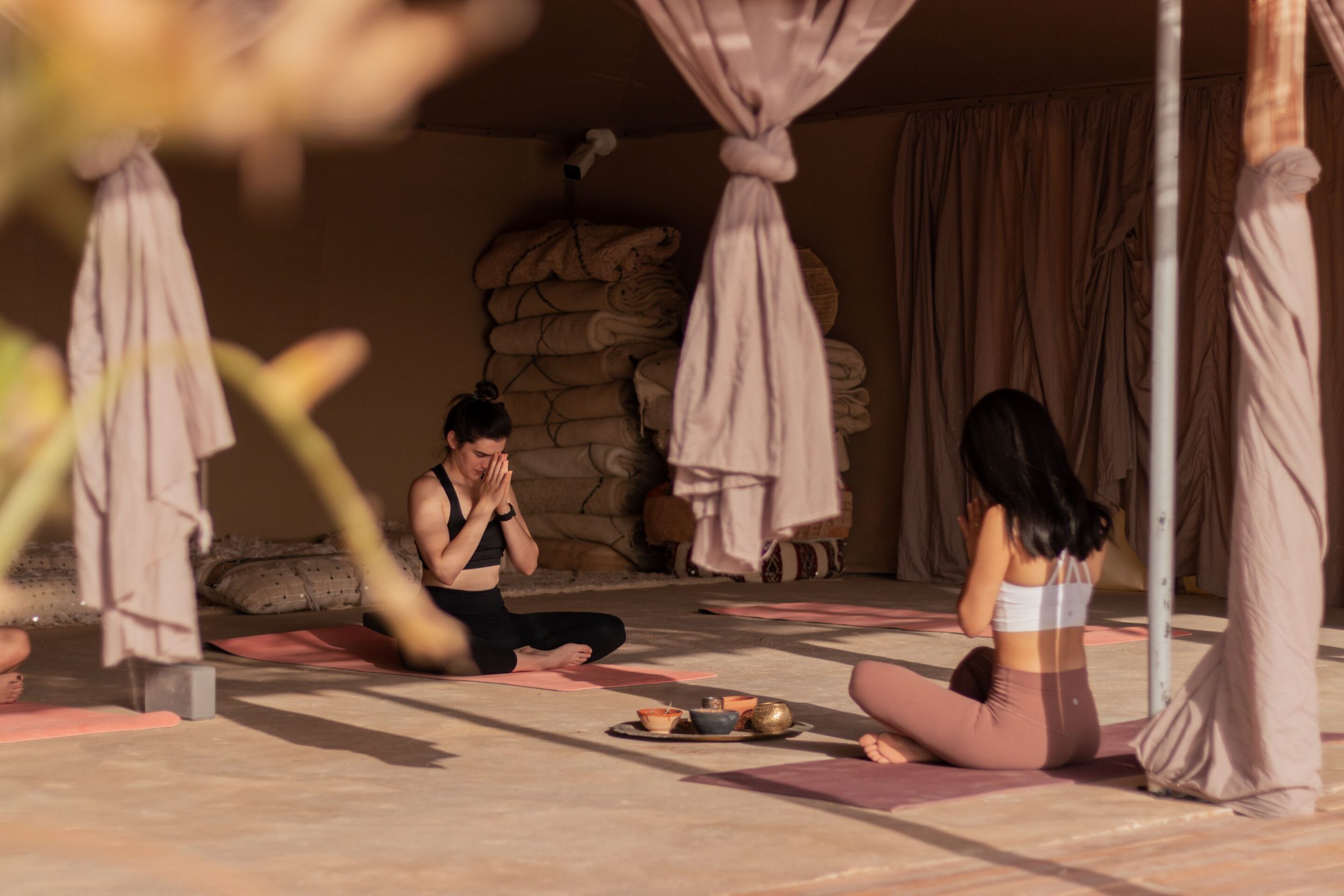 Two women sitting in meditation on yoga mats inside a desert camp tent in the Agafay, with Moroccan rugs and a tea tray