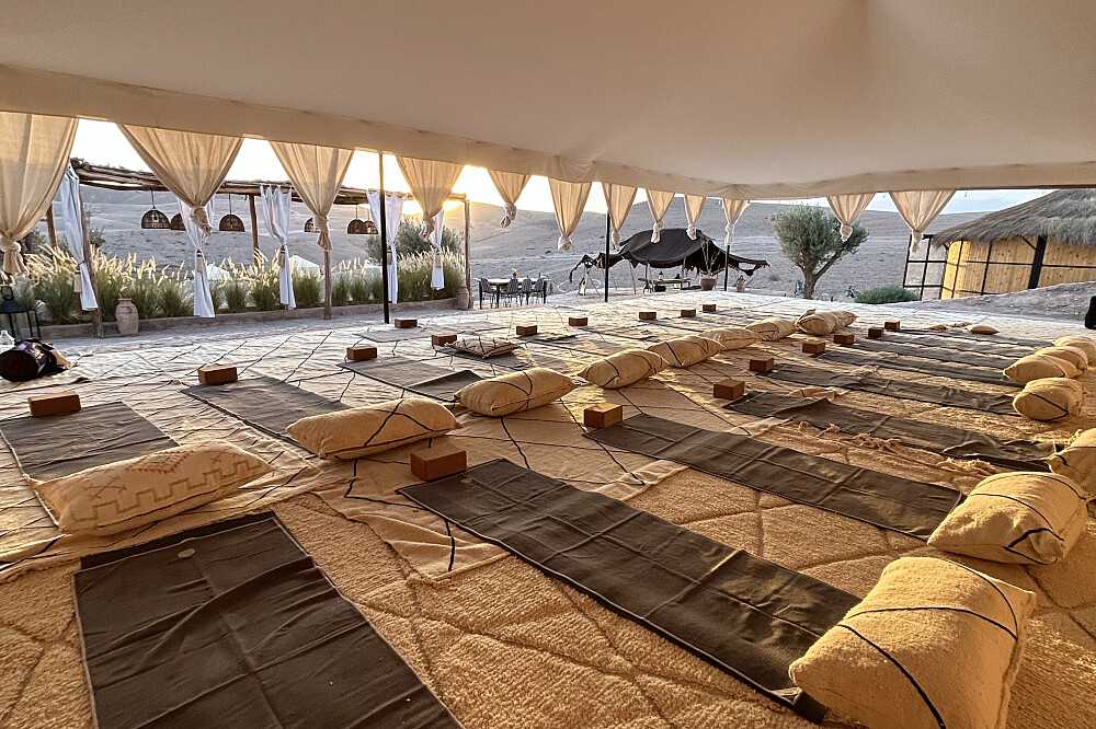 Open tent in de Agafay-woestijn met yogamatten en kussens klaargelegd, uitkijkend over de stenen vlakte bij zonsondergang