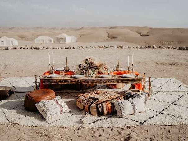 Chic picnic table set in the Agafay desert with rugs, poufs and desert camps behind